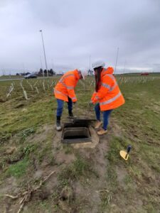 Two individuals in safety gear examining an open underground utility box in a field.