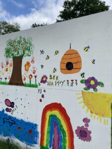 A colourful mural painted on an outdoor wall. The mural features a tree with a brown trunk and green leaves made from handprints. To the right, there’s a depiction of a bright yellow sun with rays and a smiling face, alongside blue clouds and raindrops. Below is green grass with flowers, one of which is being watered by a watering can. On the left side, there’s an image of a fish, next to a vibrant rainbow arching over the grass. 