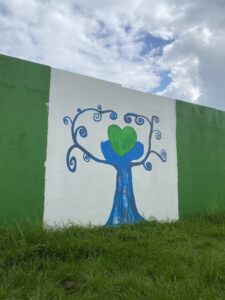 A colourful mural painted on an outdoor fence at the Brighouse Flood Alleviation Scheme. The mural features a tree with a blue trunk and a green heart. The sky in the background is partly cloudy.