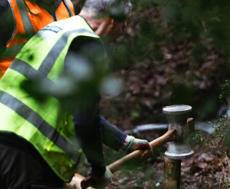 Two Slow the Flow volunteers with their faces obscured are engaged in outdoor conservation work. One is wearing a high-visibility vest and is using a tool to work on a wooden post that is part of an environmental project in a wooded area. Slow the Flow aims to reduce the impact of flooding through natural flood management techniques.
