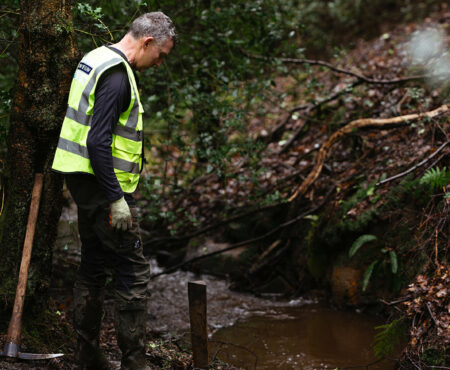 A Slow the Flow volunteer in a high-visibility vest stands beside a small stream in a wooded area, looking down at the water. There is a muddy bank with fallen branches around the stream. The setting appears to be natural and somewhat untamed.
