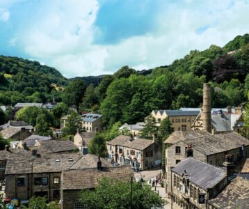An elevated view of a historic village nestled in a valley, featuring traditional stone buildings with grey slate roofs. A tall, stone industrial chimney stands among the houses and lush green trees. In the background, steep, wooded hills rise toward a bright sky with scattered white clouds.