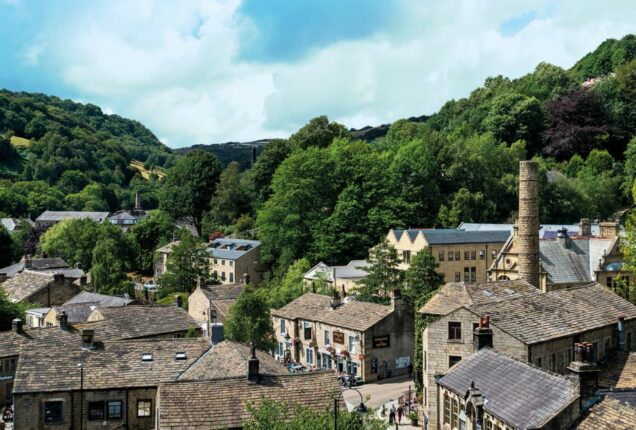 An elevated view of a historic village nestled in a valley, featuring traditional stone buildings with grey slate roofs. A tall, stone industrial chimney stands among the houses and lush green trees. In the background, steep, wooded hills rise toward a bright sky with scattered white clouds.
