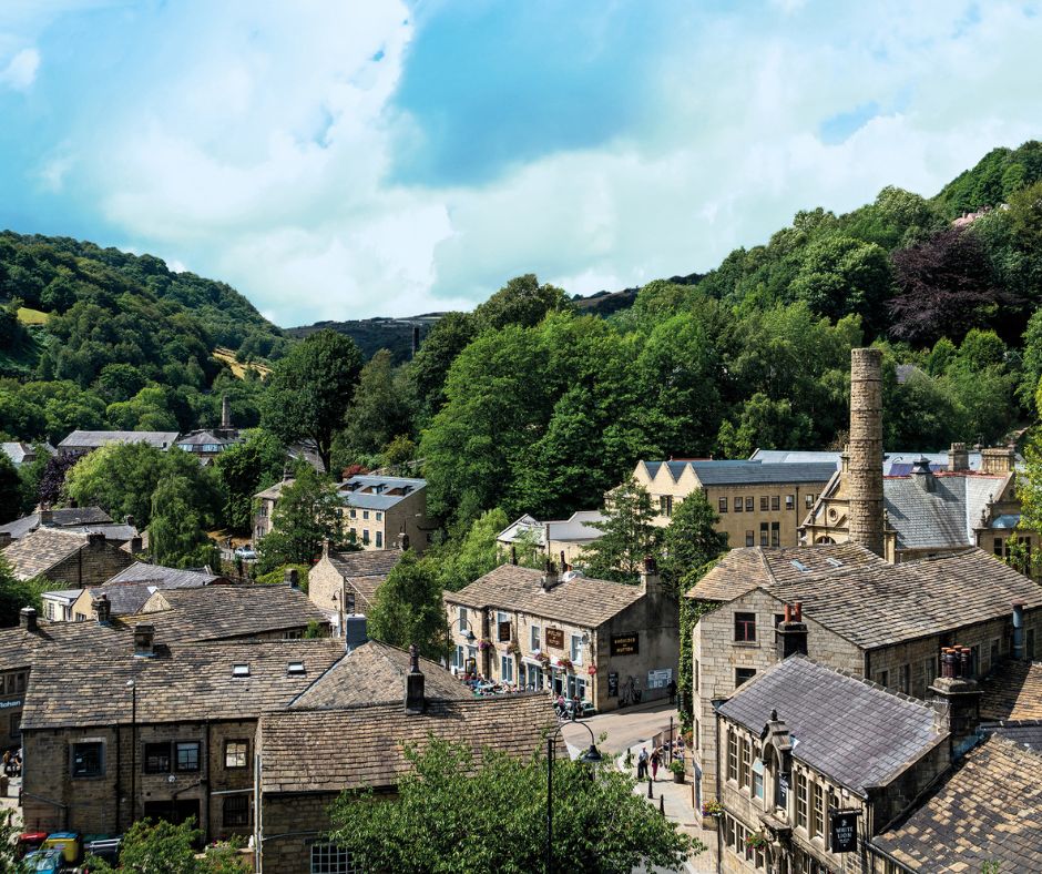 An elevated view of a historic village nestled in a valley, featuring traditional stone buildings with grey slate roofs. A tall, stone industrial chimney stands among the houses and lush green trees. In the background, steep, wooded hills rise toward a bright sky with scattered white clouds.