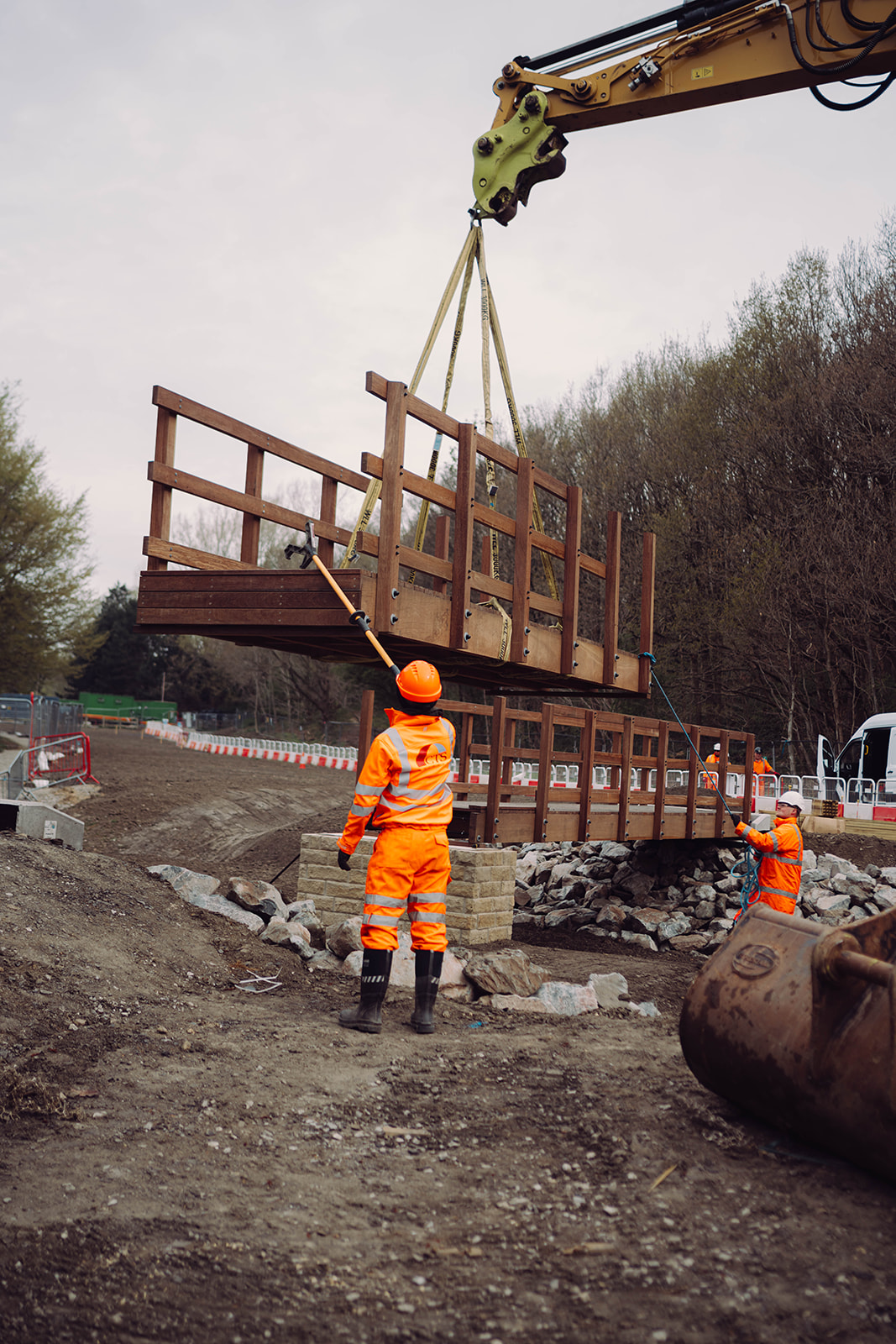 Brighouse Flood Alleviation Scheme at Wellholme Park. Image shows large machinery lowing a section of bridge into place. People working on the bridge are wearing orange high visibility clothing and hard hats. The scene shows large boulders and trees surround.