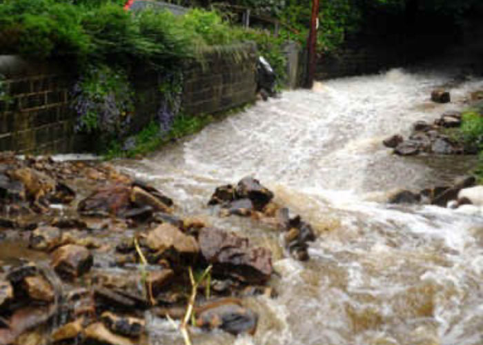 Localised flash flooding. Fast‑flowing floodwater rushing over rocks beside a stone wall.