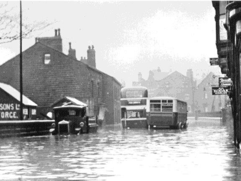 Mytholmroyd bus flooded in 1950s. Old buses partly submerged in deep floodwater on a street. Nearby buildings and road signs are also surrounded by floodwater.