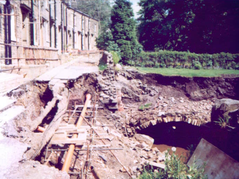 Damage caused by flooding in 1980s in Calderdale. Severe ground erosion beside a row of stone houses, exposing pipes and a collapsed section near an old brick archway.