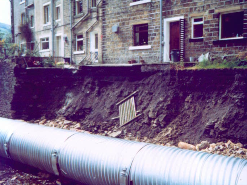 Damage caused by flooding in 1980s in Calderdale. Large metal drainage pipe installed below an eroded embankment behind a row of stone buildings.