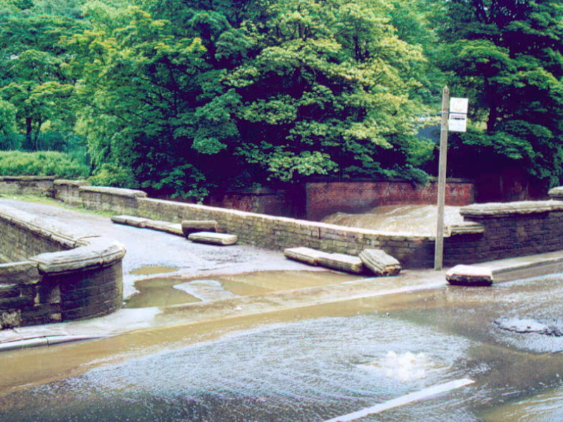 Floodwater flowing across a stone bridge and nearby road in a tree-lined area.