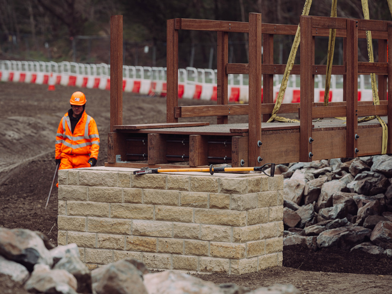 Brighouse Flood Alleviation Scheme bridge installation. Construction worker beside a partially installed wooden footbridge on a stone base.