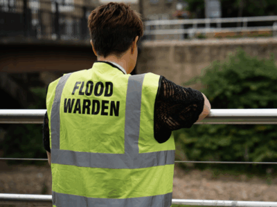 Person wearing a high‑visibility vest labeled “FLOOD WARDEN,” standing by a railing near a river.