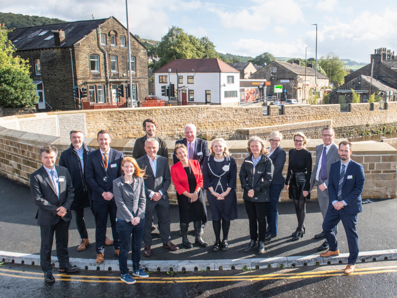 Gemini said An outdoor group portrait of thirteen people standing on a paved road in front of a newly constructed stone flood wall. The group, a mix of men and women dressed in professional and business-casual attire, is positioned in two rows. In the center of the group, Tracy Brabin, the Mayor of West Yorkshire, stands smiling in a dark dress with white trim. Behind the group, a curved, light-brown stone wall follows the line of a river. In the background, traditional stone buildings, a "Sainsbury's Local" storefront, and green hills under a bright, slightly hazy sky are visible. The scene is lit by clear, direct sunlight.