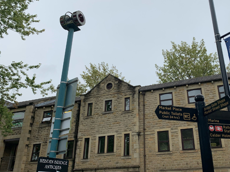 Hebden Bridge flood siren shown to the backdrop of buildings in the centre of Hebden Bridge. The image also shows street signs for local attractions in Hebden Bridge, such as Market Place.