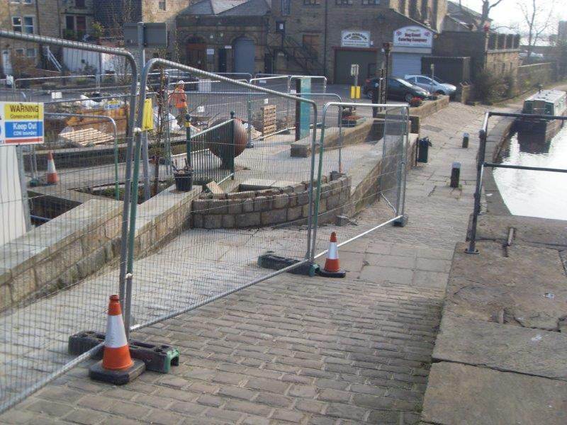 Flood alleviation work on a wall in Todmorden. Temporary fencing and traffic cones cordoning off a construction area beside a canal walkway.