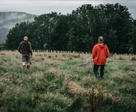 Two people walking through a grassy field with tall, wild grass. Dense trees and misty rolling hills appear in the background under an overcast sky.