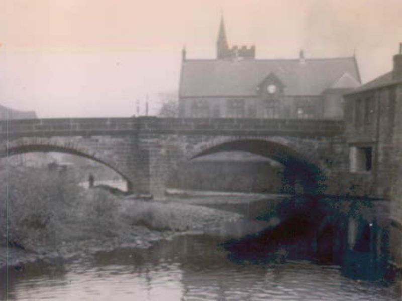 An old sepia-toned photograph capturing a tranquil riverside scene. In the foreground, a gently flowing river. A stone bridge spans the water, leading the eye toward a historic church with a tall spire in the background.