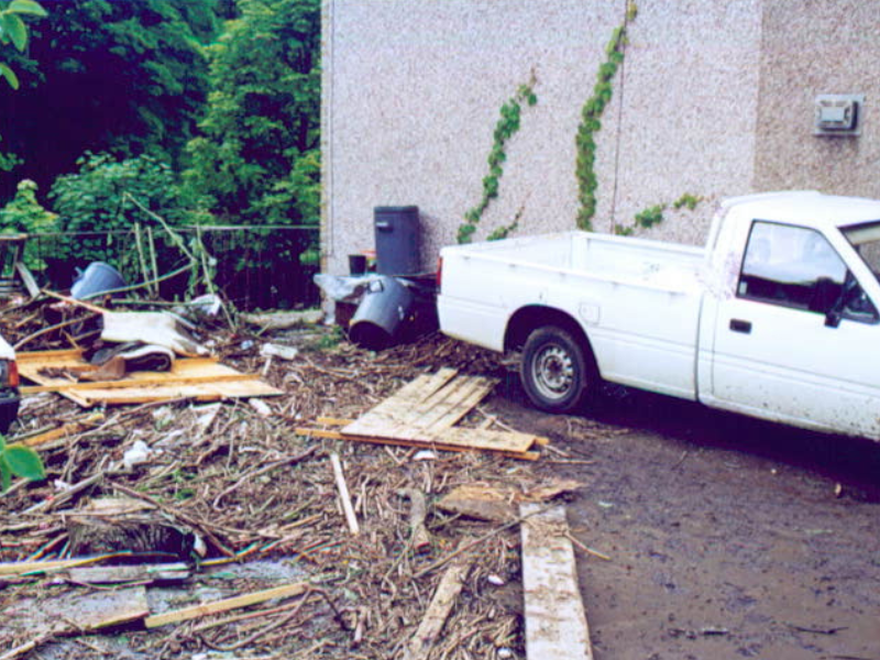 Aftermath of a flood showing a residential driveway littered with debris. Broken wooden fences, branches, and other scattered materials block the entrance, with muddy water stains visible on the ground. The scene reflects the damage and disruption caused by the flooding.