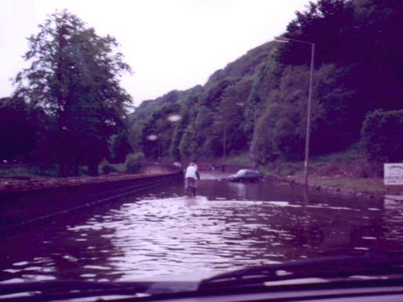 An old photo that shows surface water flooding on a road in Calderdale. The picture is taken from a car, and you can see a cyclist navigating through the floodwaters, along with a couple of abandoned cars at the far end of the road.