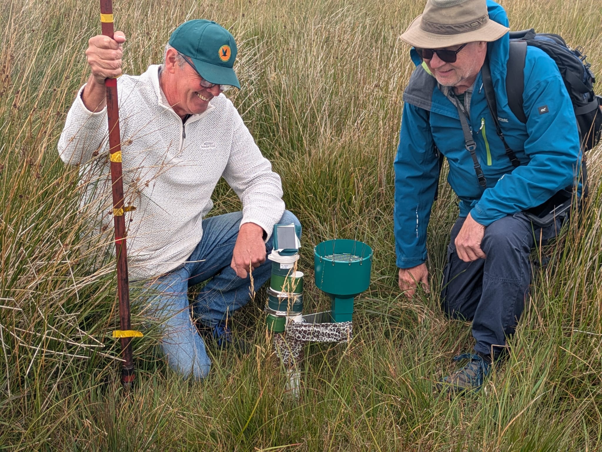 wo volunteers working beside a rain gauge at Bridestones, kneeling in long, dense moorland grass. The rain gauge sits low to the ground and includes a cylindrical green collection funnel and attached monitoring components. One volunteer holds a survey pole marked with measurement bands, and both are wearing outdoor clothing suitable for fieldwork. The surrounding landscape is open moorland with tall grasses under an overcast sky.