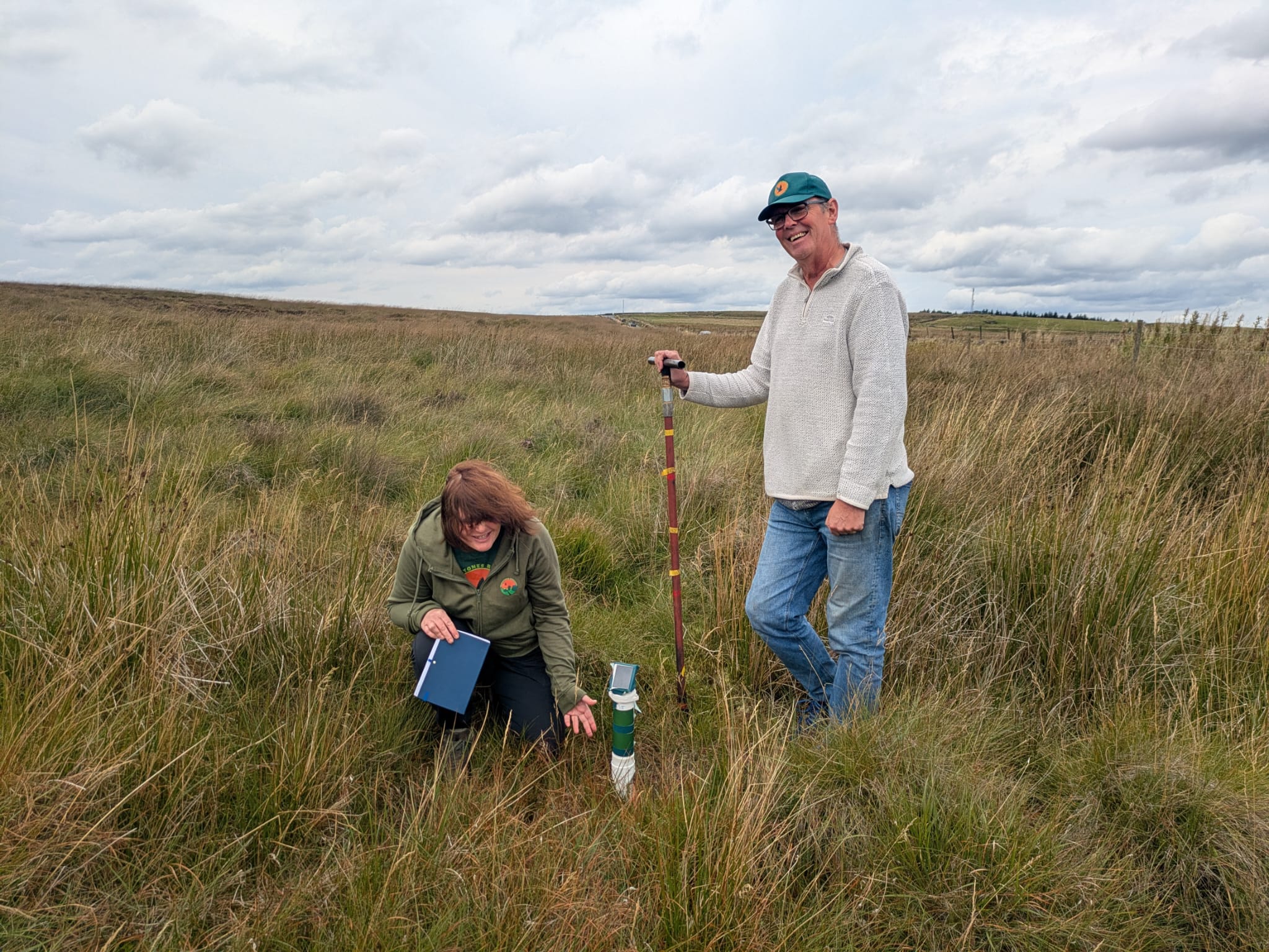 Two volunteers in an open moorland landscape standing beside a flow monitor at Bridestones. One volunteer is kneeling in long grass with a notebook, while the other stands holding a survey pole next to the monitoring device. The scene is surrounded by tall grasses under an overcast sky.