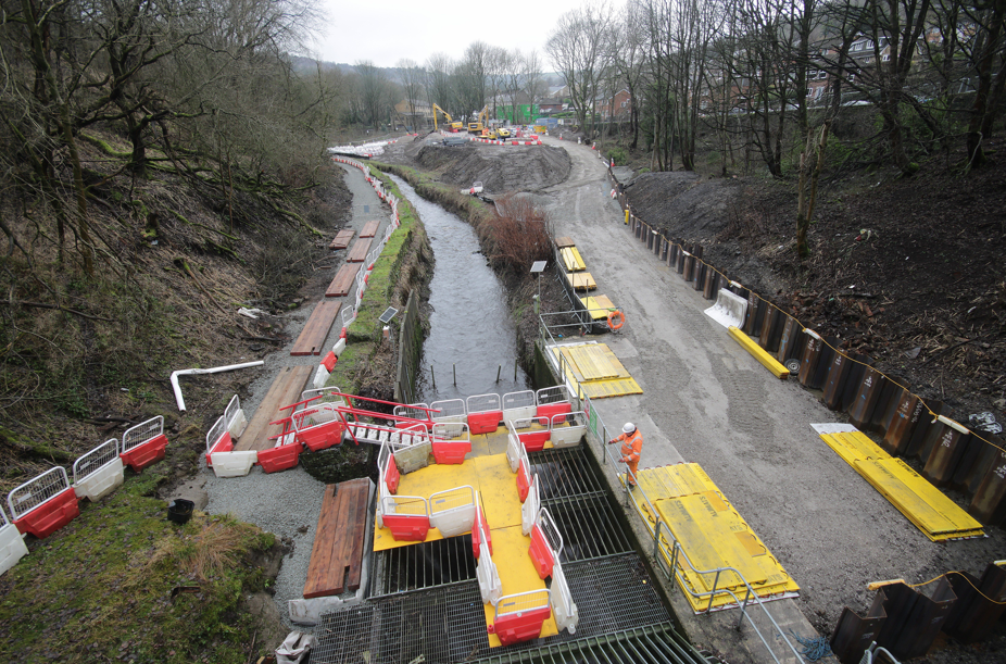 Construction work at Hebble Brook. onstruction works along a riverside channel with barriers, walkways and heavy machinery, as workers carry out flood resilience works in a wooded area.