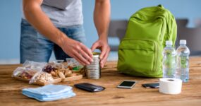 Hands are placing a metal can into a green backpack on a table full of emergency supplies like food, water, and personal items.