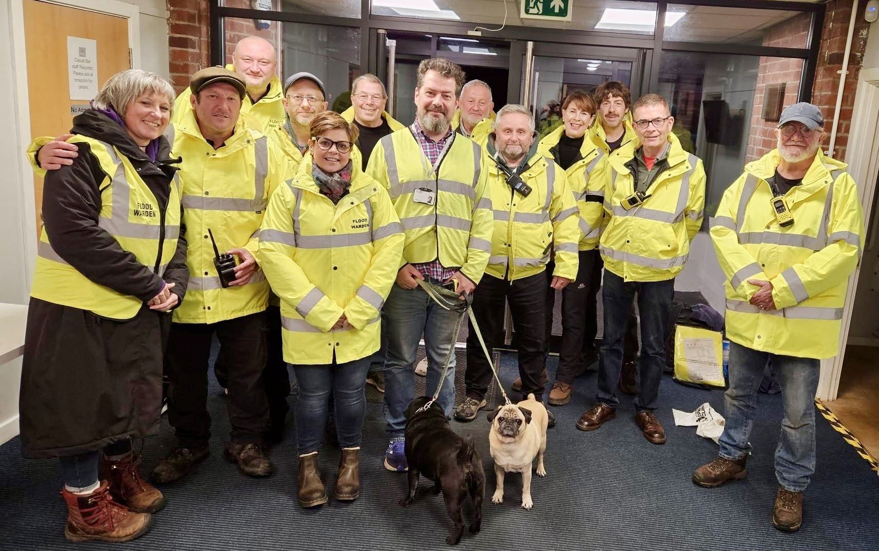 A group of about twelve people stand together indoors, posing for a group photo. They are all wearing bright neon yellow high-visibility jackets, some with "FLOOD WARDEN" printed on them. A few individuals also have walkie-talkies attached to their clothing. In the front center, a man holds the leashes of two pugs—one black and one fawn—standing on the floor. The background shows a doorway and a brick wall.