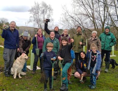 A group of eleven smiling volunteers of various ages pose together outdoors on a grassy field, holding shovels and tools for a tree planting event. A golden retriever sits with them, and newly planted saplings in protective guards are visible in the background.