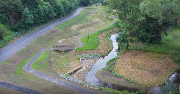 An aerial view of Wellholme Park featuring a winding stream on the right, a wooden footbridge spanning over a dry ditch, and a paved walking path running parallel to a dense line of trees on the left. The landscape includes grassy banks, rocky stream edges, and a winding asphalt path cutting through the green space.