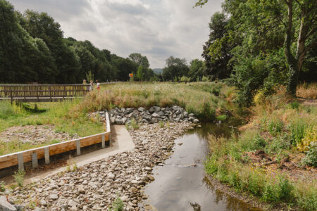 A wide view of new flood defence infrastructure along the stream at Wellholme Park, featuring stone riverbank reinforcement, a wooden barrier wall, a footbridge, and surrounding natural greenery.