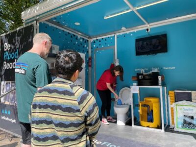 A flood resilience staff member in a red shirt demonstrating property flood protection equipment inside the mobile Flood Pod trailer to two visitors outside.
