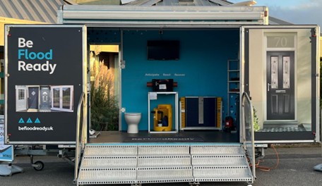 Straight-on view of the mobile Flood Pod trailer fully open, featuring prominent "Be Flood Ready" branding and displaying property resilience measures like flood doors, barriers, and pumps inside.