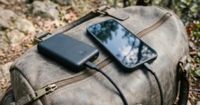 A portable power bank charges a black smartphone resting on top of a packed emergency bag, illustrating the importance of staying connected and powered during emergencies such as flooding.