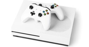 White games console with two white wireless controllers resting on top, photographed against a light grey background. The controllers feature black thumbsticks and colourful action buttons, while the console has a perforated ventilation pattern on one side.